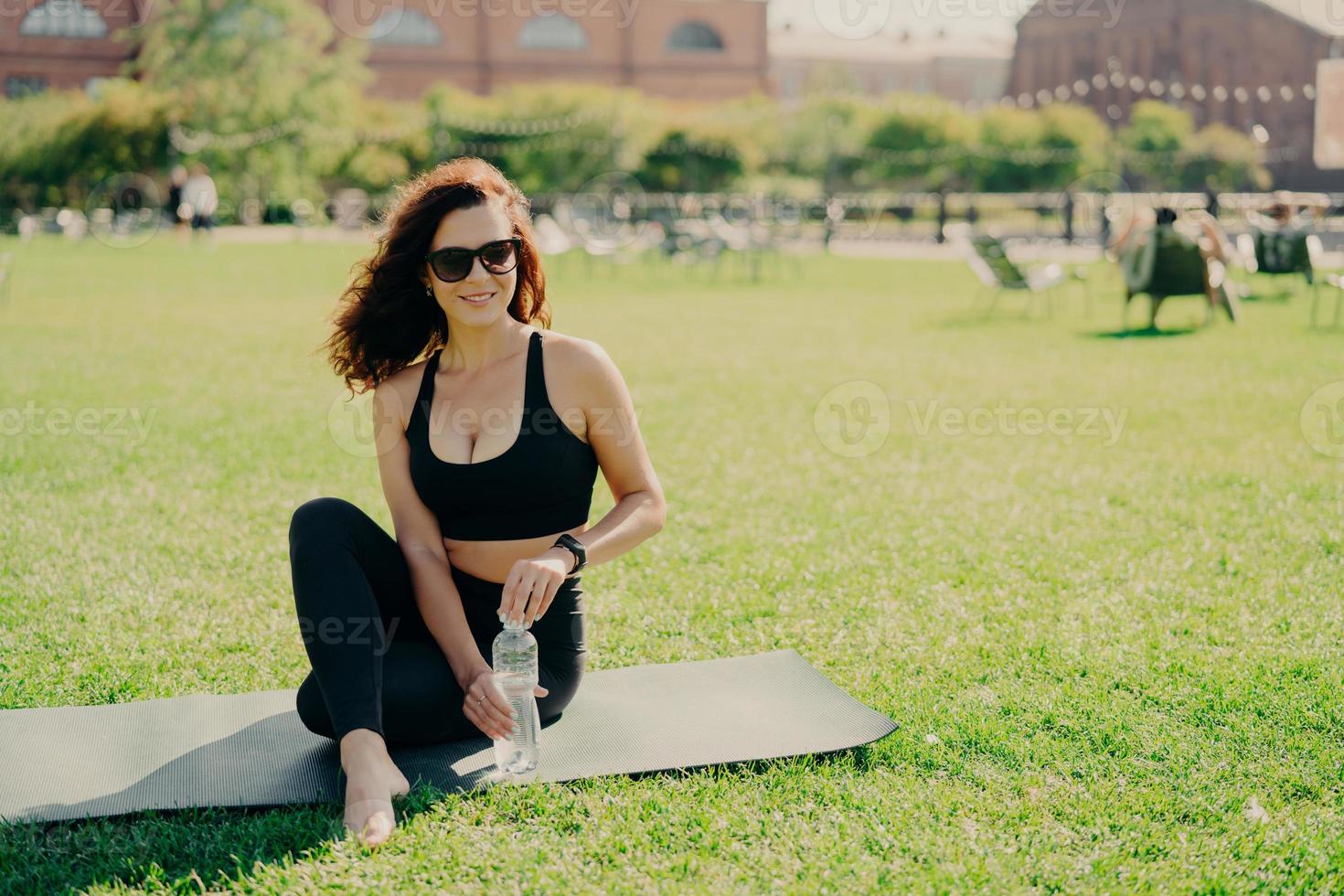 Horizontal shot of active slim woman in sportswear sits on karemat holds bottle of cold water has rest after exercising abdominal muscles poses on green lawn wears sunglasses. Workout at street photo