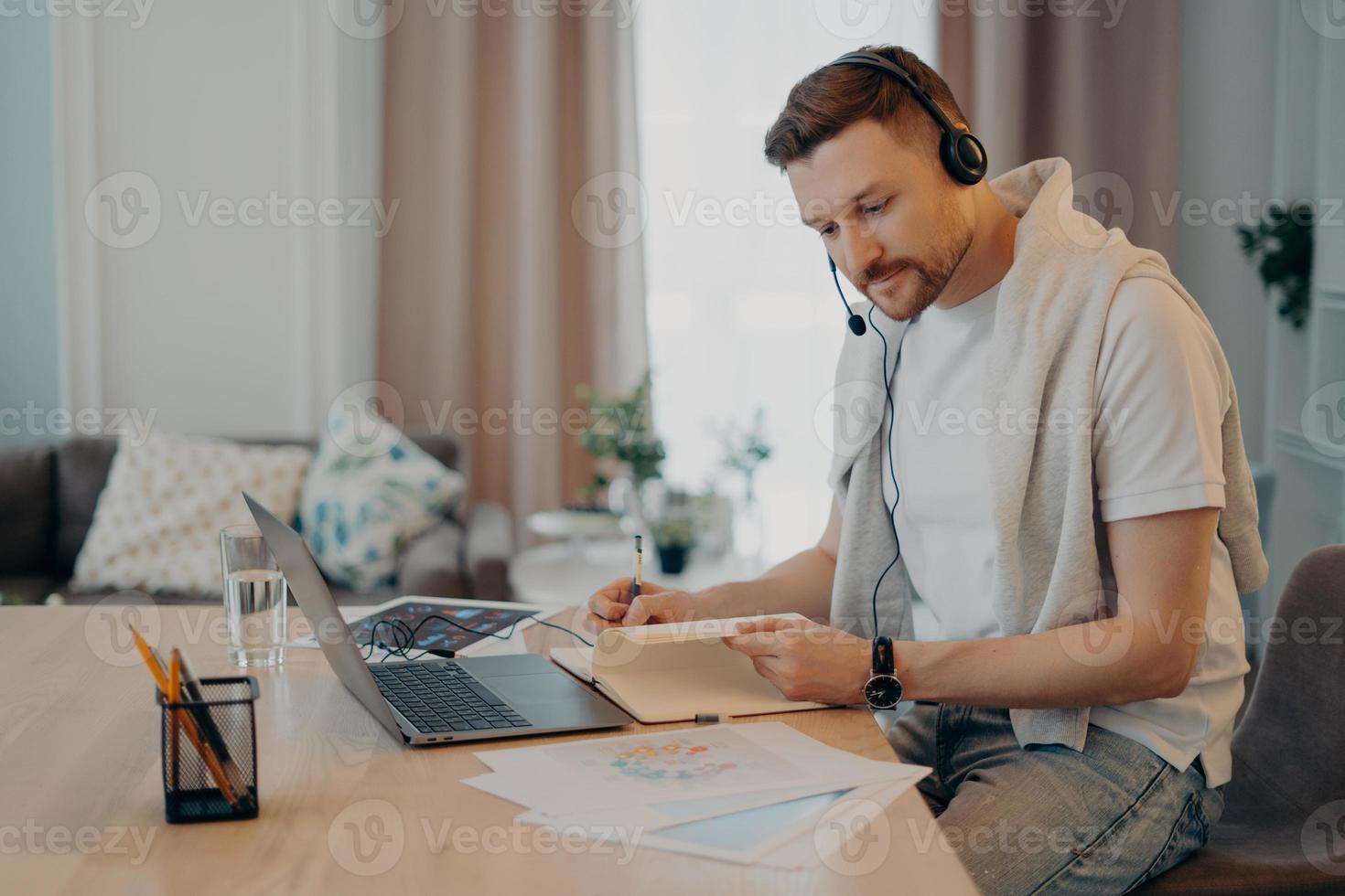 Focused bearded guy using laptop and notebook while working remotely photo