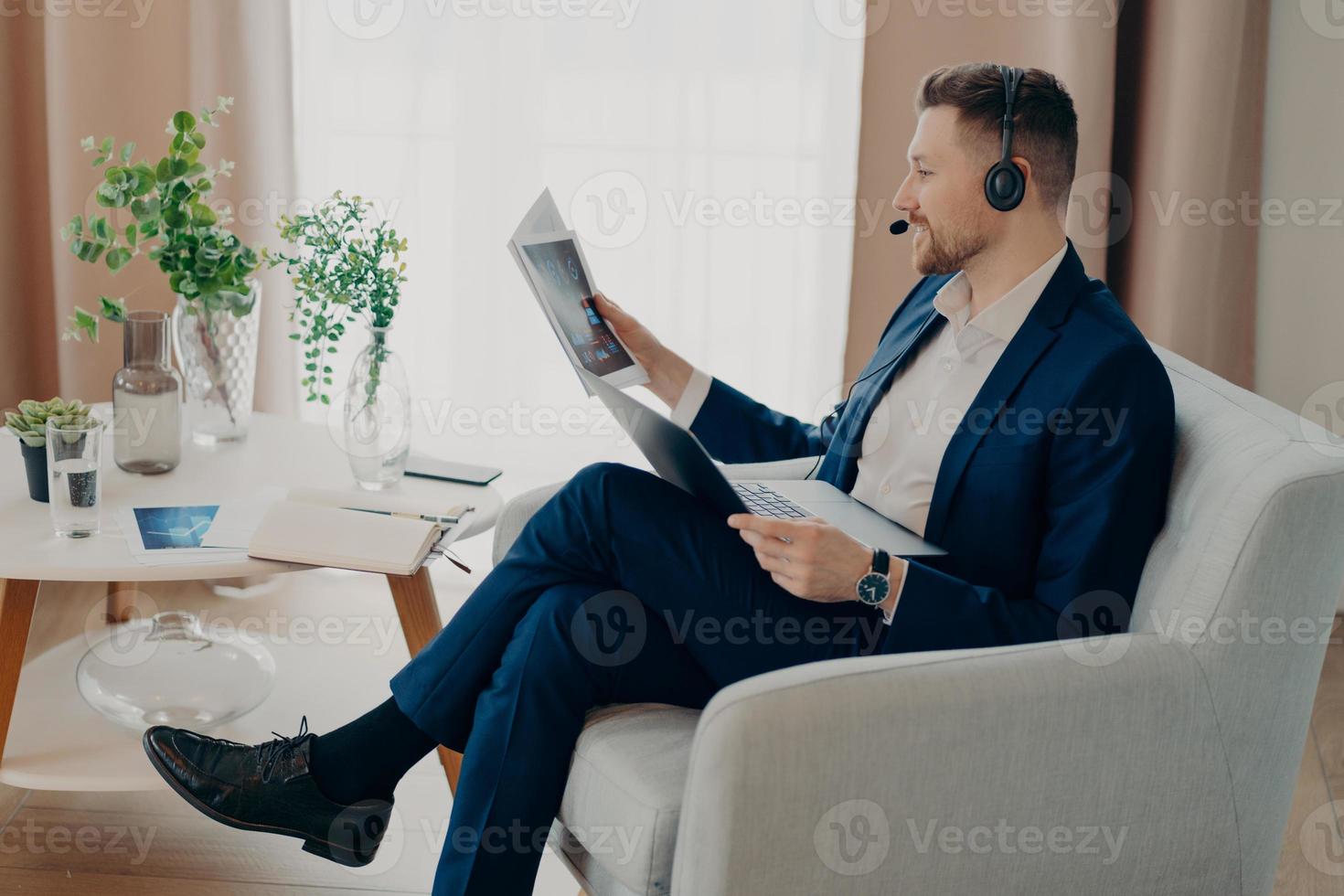 Sideways shot of bearded male corporate worker analyzes data looks through documents with graphics reviews financial report dressed in formal clothes poses in modern apartment uses laptop headset photo