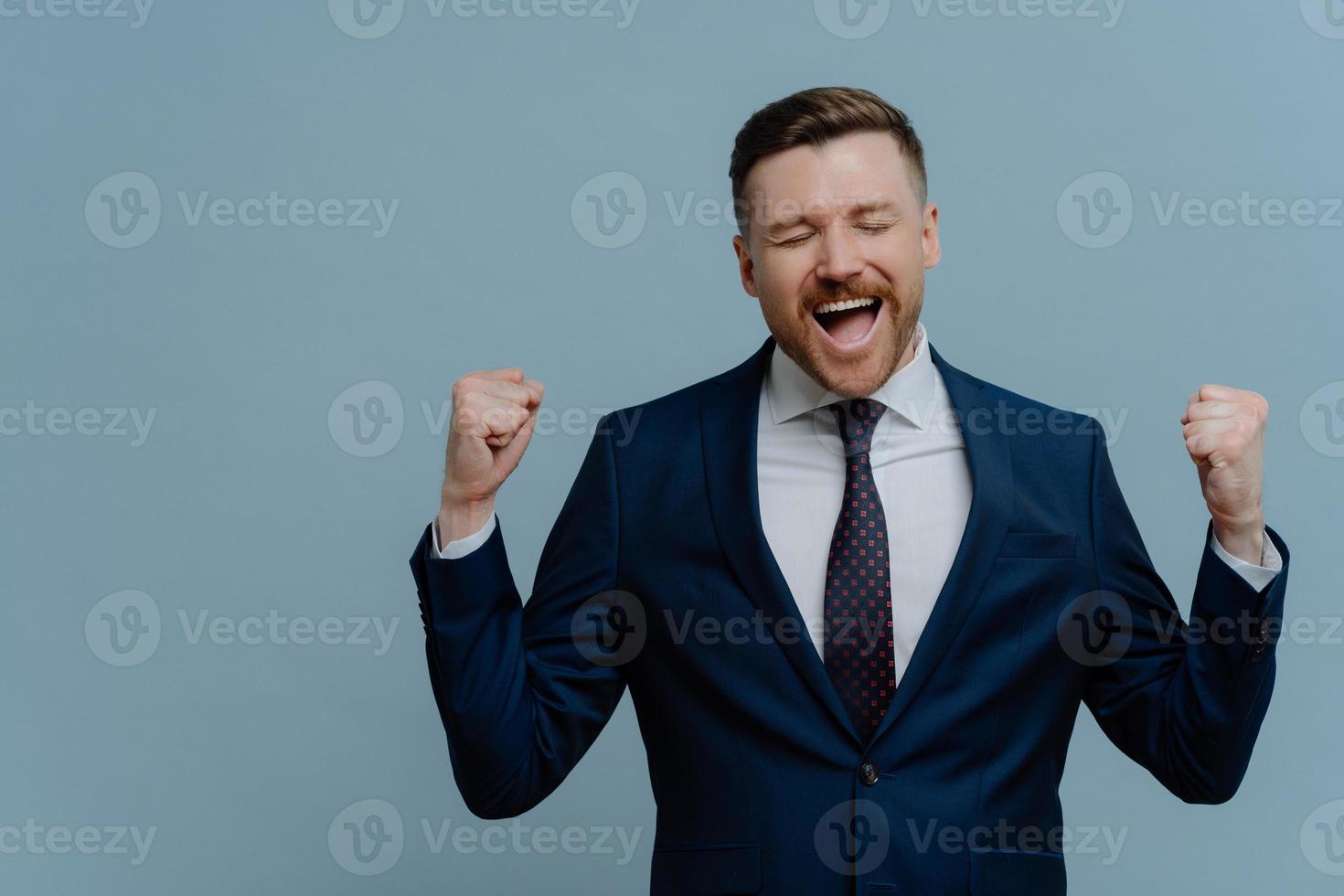 Overjoyed triumphing businessman clenches fists with triumph celebrates successful deal dressed in formal clothes isolated over blue background copy space for your advertisement exclaims from joy photo