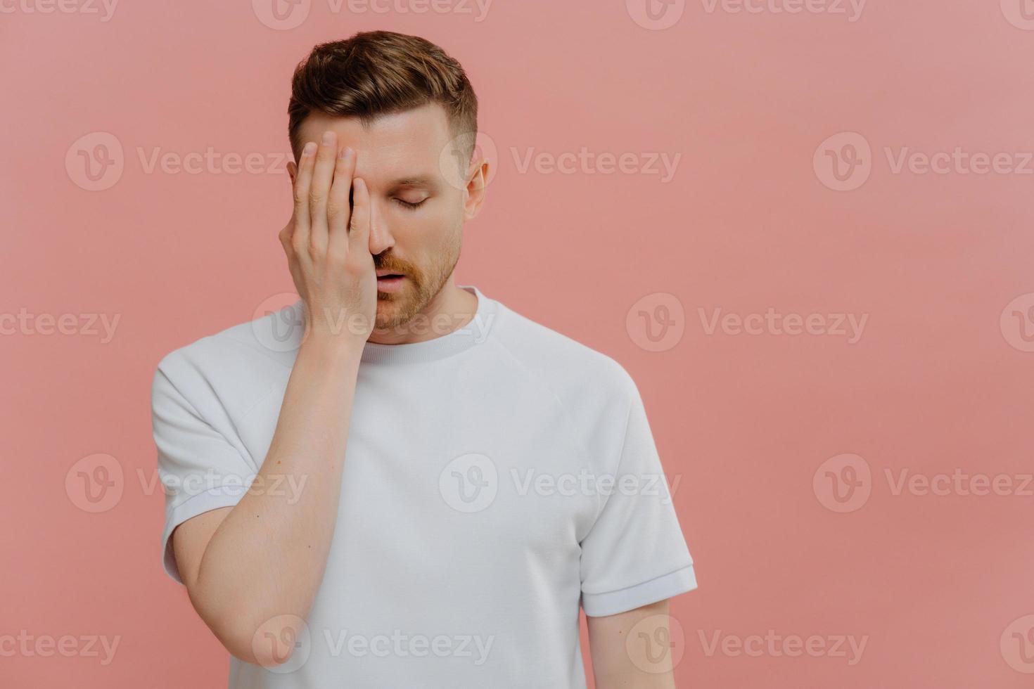 Horizontal shot of serious tired man makes face palm feels bored spending time alone covers eye with hand dressed in casual white t shirt isolated over pink background blank space on right side photo