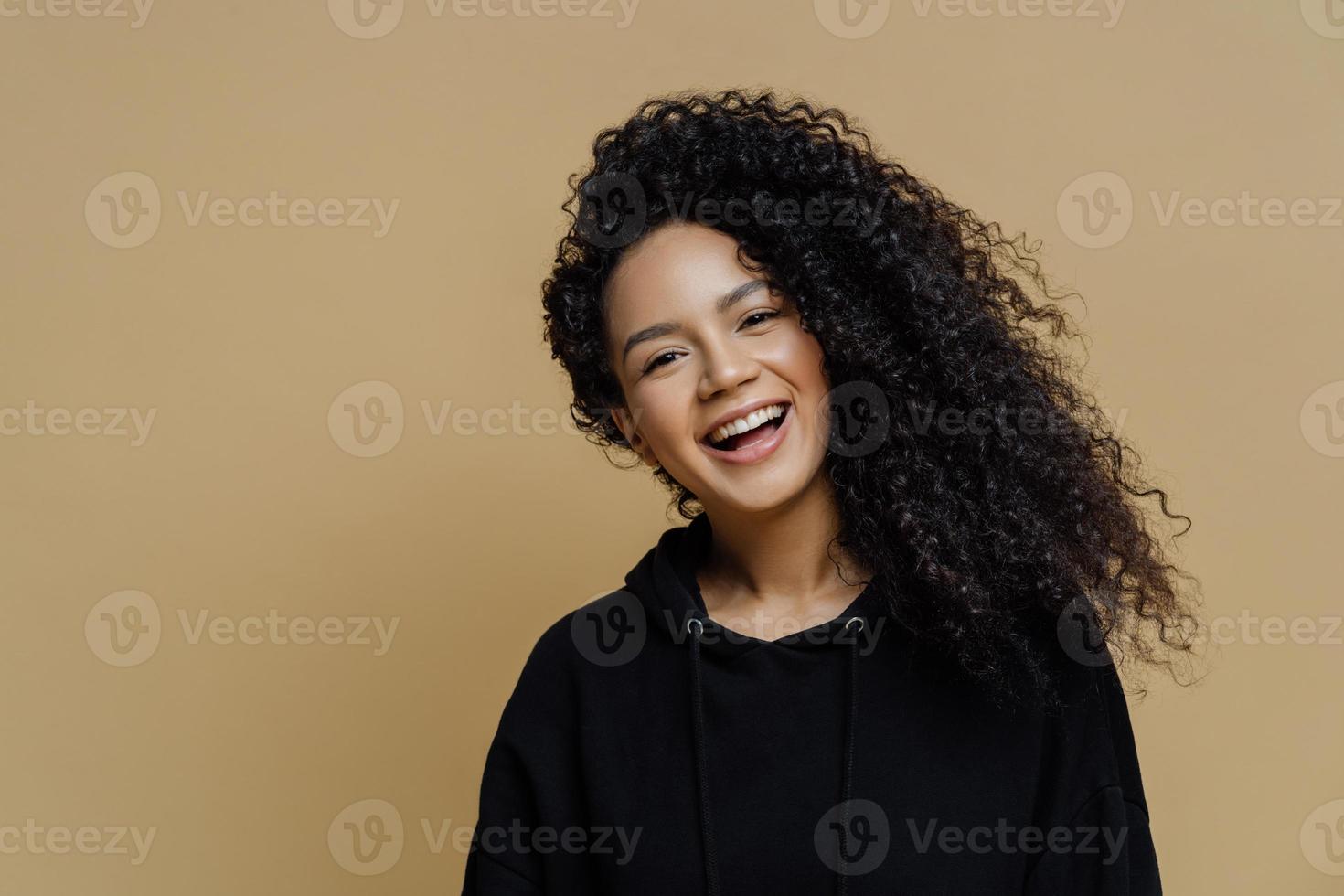 feliz mujer afroamericana positiva con cabello rizado, inclina la cabeza y sonríe ampliamente, vestida con una sudadera negra informal, aislada en un fondo beige, expresa emociones positivas. gente, felicidad foto