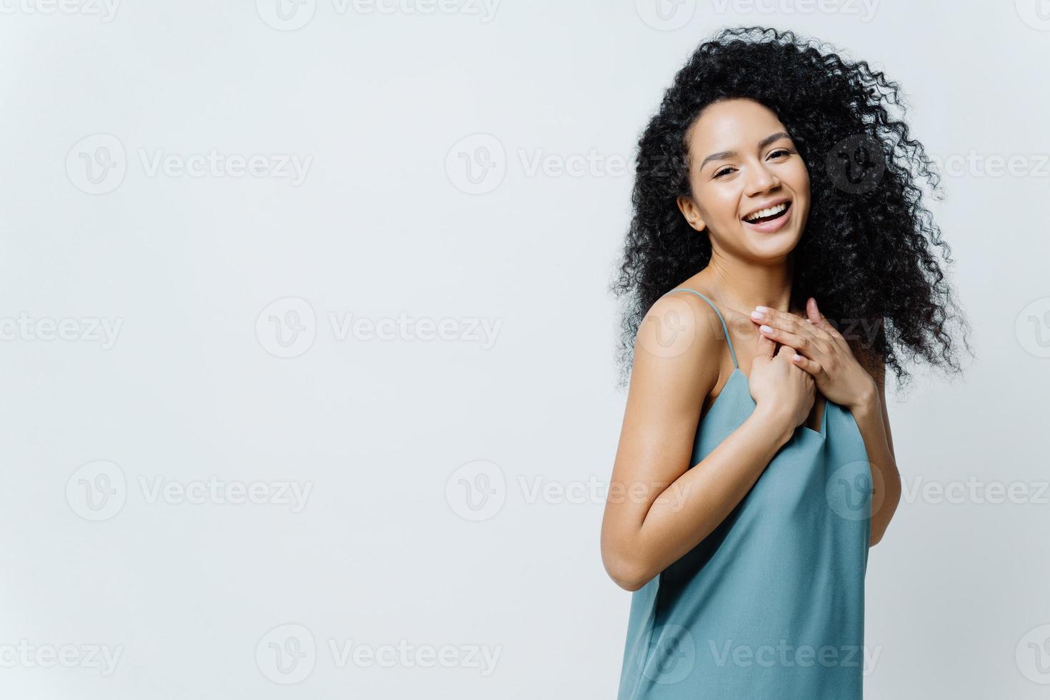 Overjoyed Afro American woman laughs positively, keeps hands on chest, hears hilarious joke, expresses happiness, wears casual dress, poses in studio against white background, giggles positively photo