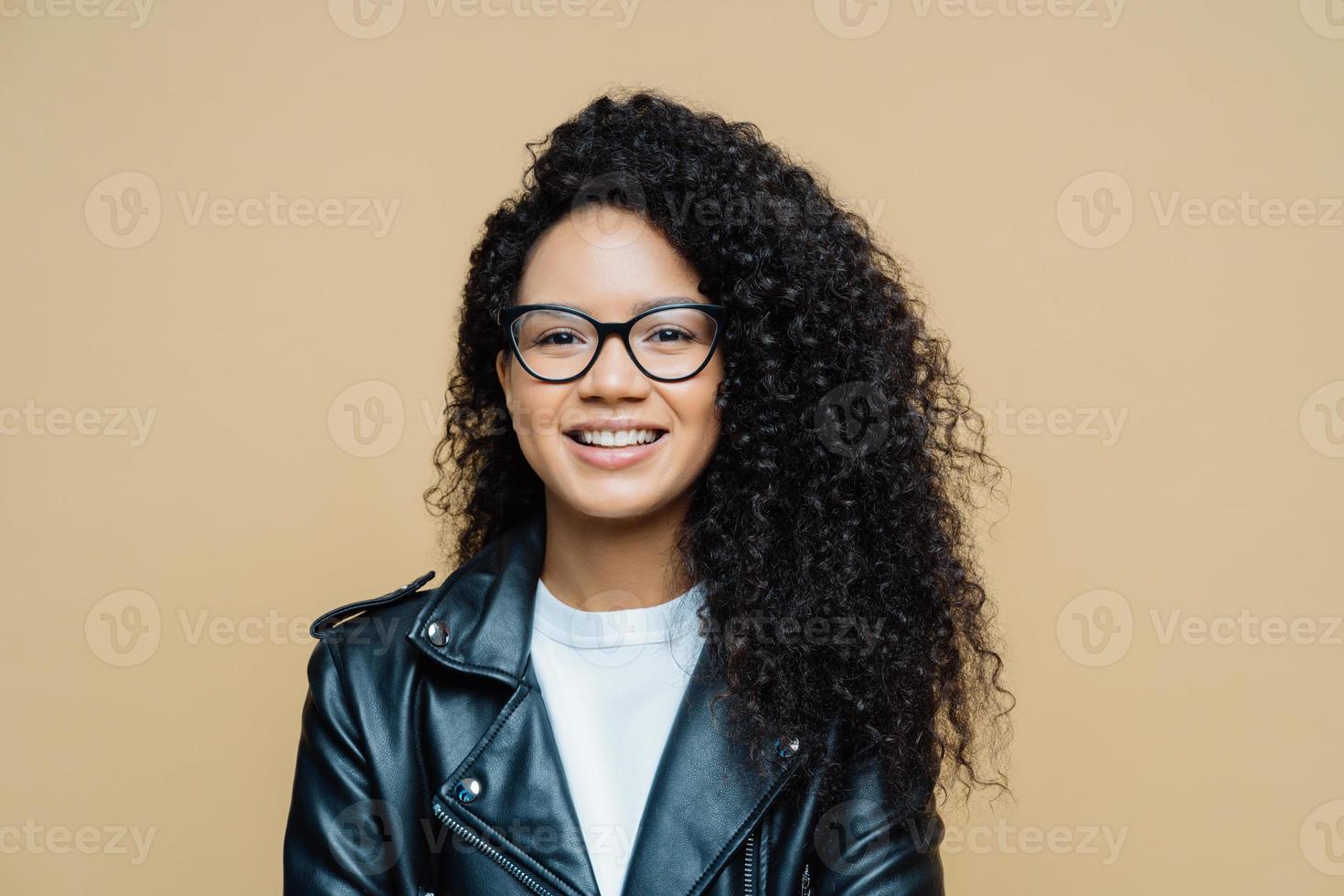 Stylish African American woman smiles toothily, has pleasant talk, enjoys good day, wears transparent glasses, leather jacket, expresses positive emotions, poses indoor at studio. Ethnicity, fashion photo