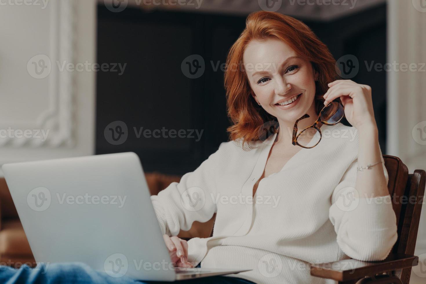 toma interior de una mujer alegre y satisfecha que trabaja de forma remota, ocupada con un trabajo a distancia, mira positivamente a la cámara antes de comenzar a trabajar, usa una computadora portátil moderna e Internet inalámbrico, busca información foto