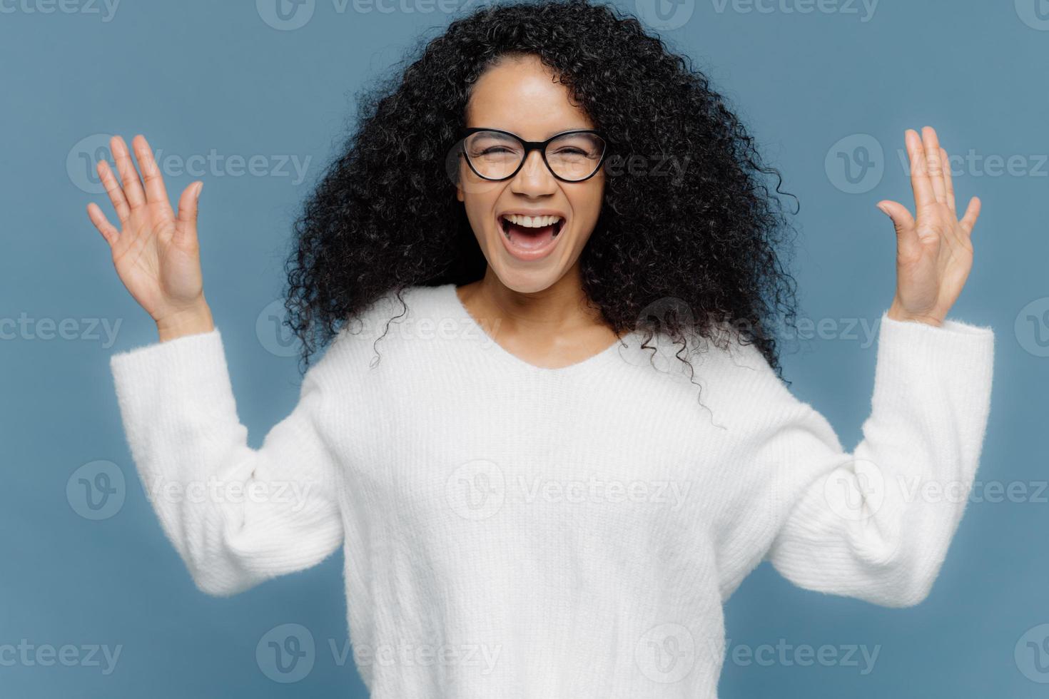 Overjoyed mixed race woman with curly hair, raises hands, exclaims from positive emotions, keeps mouth opened, dressed in white sweater, stands against blue background. Facial expressions concept photo