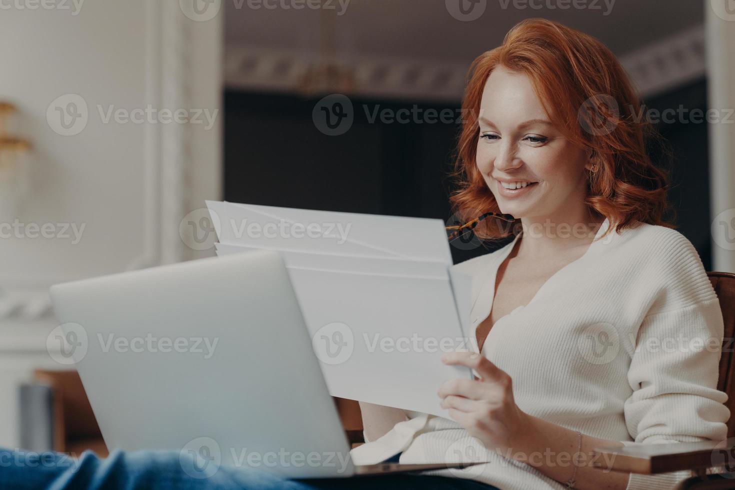 Horizontal view of pleased adult woman entrepreneur works from home, works remotely from home, looks through paper documents, uses laptop computer, wears casual jumper, poses in own cabinet. photo