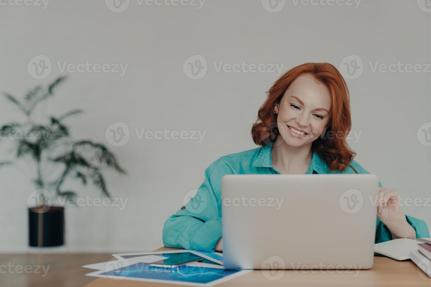 Horizontal shot of happy ginger woman freelancer works on laptop computer at home, studies online, uses software, surfs information in internet, sits at desktop, smiles positively, works remotely photo