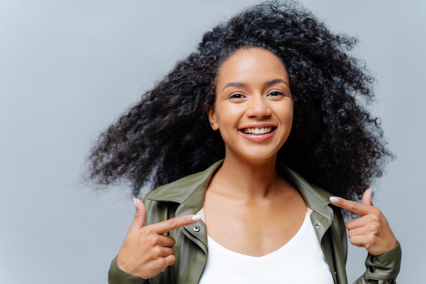 Headshot of pretty smiling woman with Afro hairstyle, points both index fingers at herself, feels proud of herself, wears leather shirt, isolated over studio background. Look at me, my new outfit photo