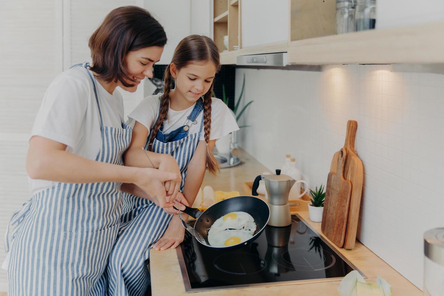 madre e hija ocupadas usan delantales a rayas, posan en la cocina cerca de la cocina, fríen huevos en la sartén, preparan un desayuno rápido, disfrutan del ambiente doméstico. mamá enseña a los niños pequeños a cocinar. concepto de familia feliz foto