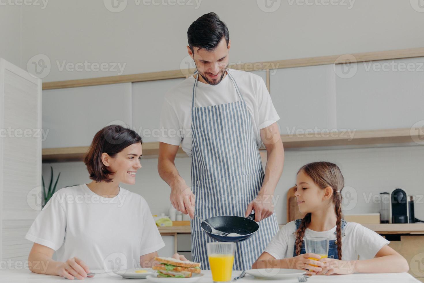 Busy caring father prepares delicious breakfast for wife and daughter, wears apron, puts fried eggs on plate. Cheerful mother and child talk to each other while sit at kitchen table, have meal photo