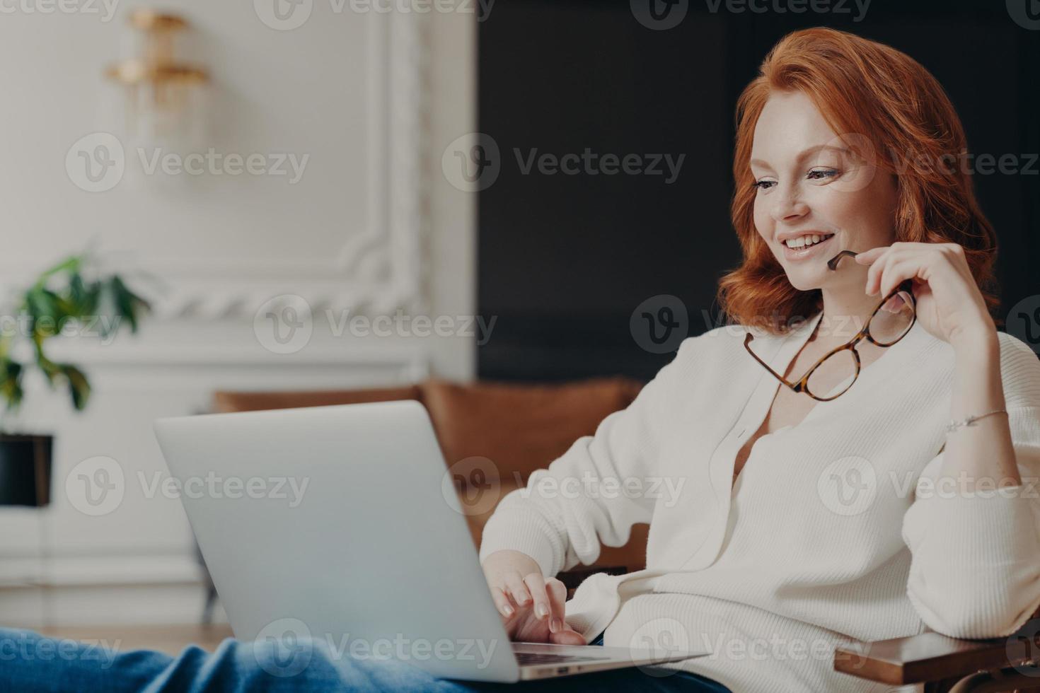 Pretty satisfied redhead female student watches tutorial video, works distantly from home, concentrated in laptop computer, prepares course work, holds spectacles, poses in modern spacious room photo