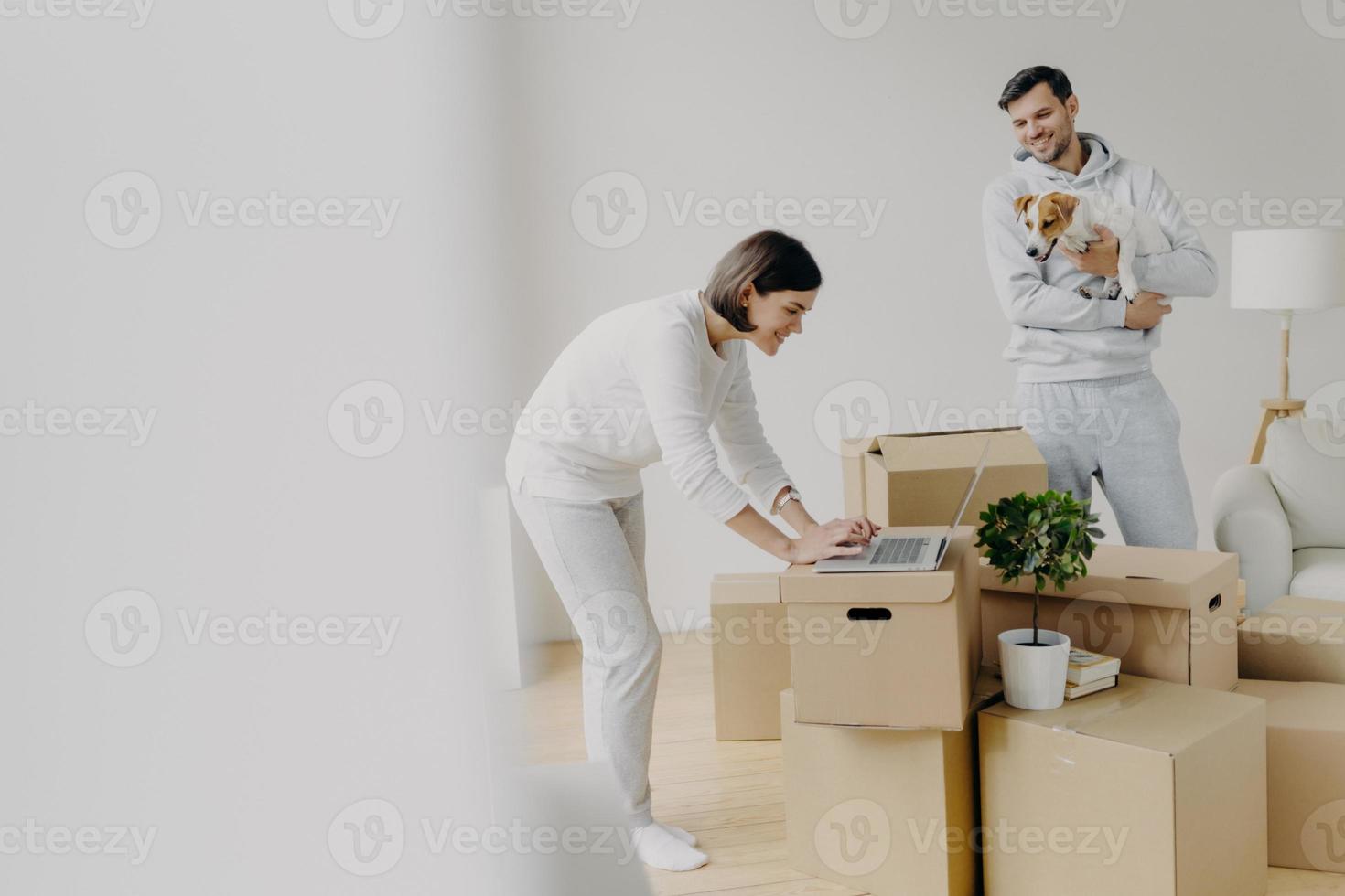 Indoor shot of attractive woman leans to laptop computer, focused into screen, surfes internet, searches ideas for decoration, man stands near, plays with favourite pet, move into new house. photo