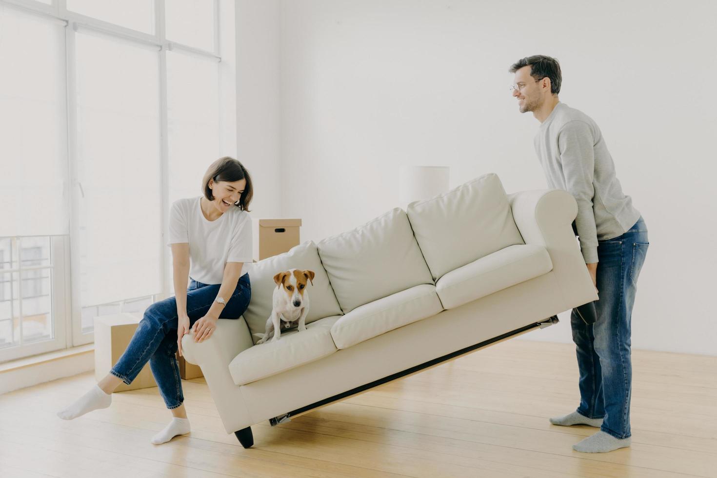 Happy couple carry modern white sofa with dog together, place furniture in living room, care about improvement of interior design, start living in new home, pose in modern apartment, have fun photo