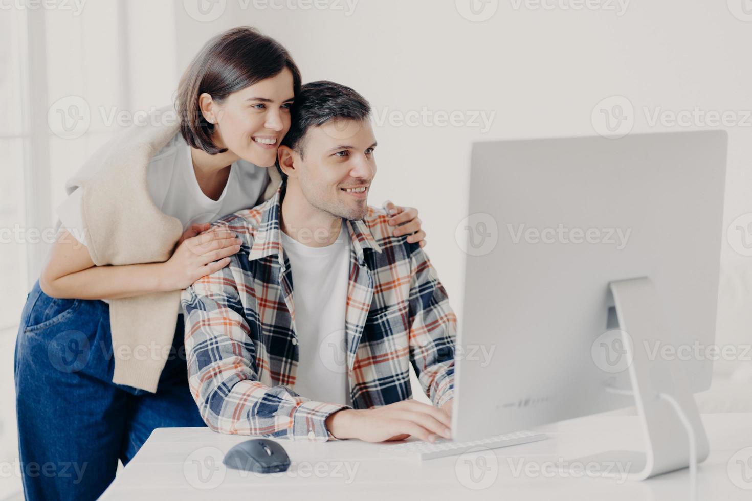 Young couple cooperate for developing website, work on common internet project, affectionate wife hugs husband with support, focused into monitor, keyboard information, connected to internet photo