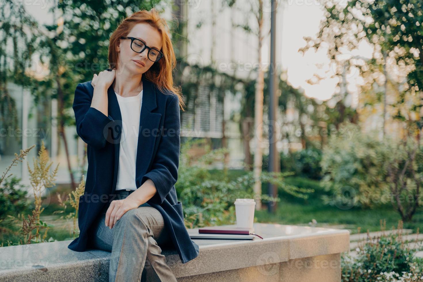 Frustrated redhead young woman feels very tired suffers from neck pain sits in park photo