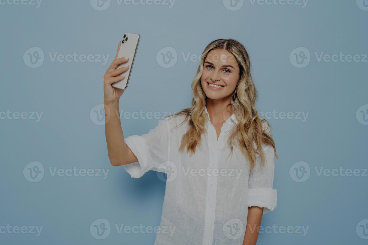 Cheerful smiling young woman with wavy long blond hair making photo on smartphone