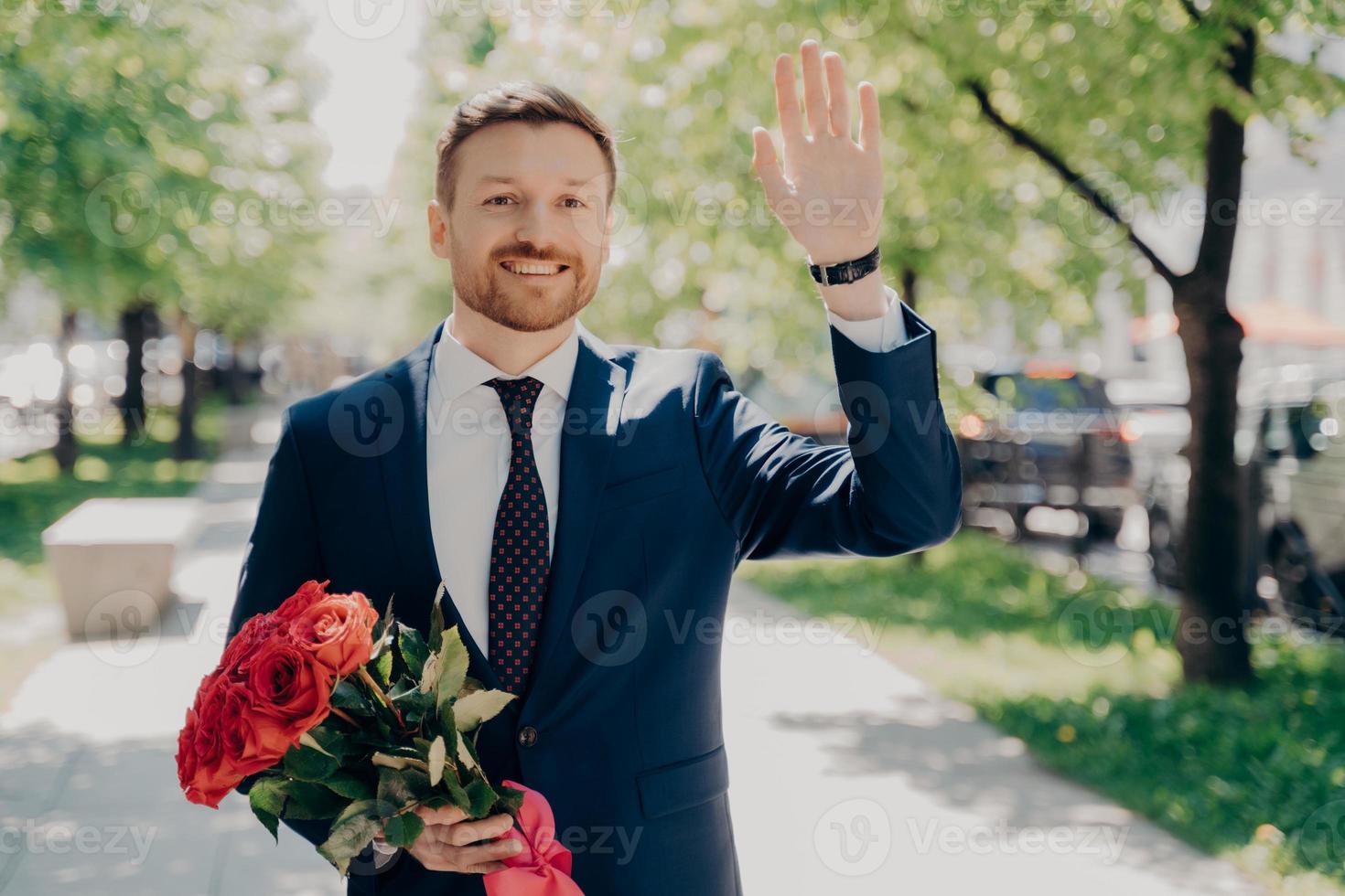 Happy handsome young man in blue suit with flower bouquet of red roses waiting for beloved woman in park photo