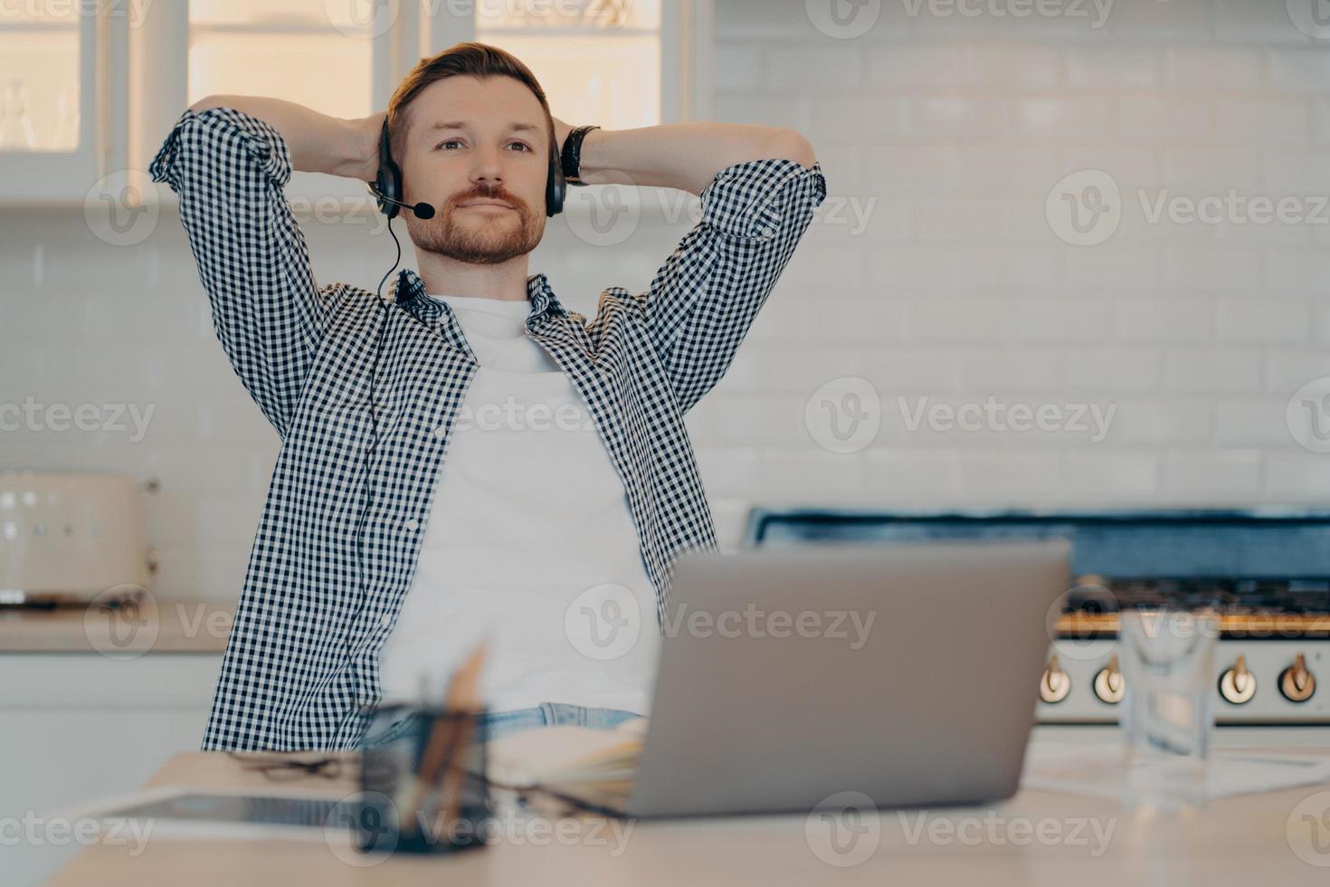 Thoughtful bearded male freelancer sits in relaxed pose takes break after working online wears headset with microphone listens educational webinar gets remote education uses new computer app photo