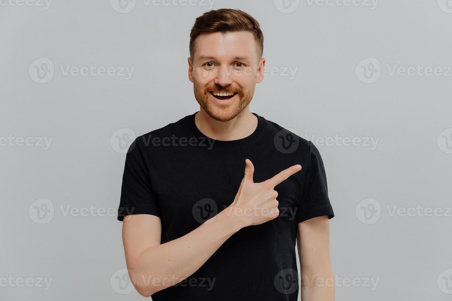 Glad handsome bearded young European man with cheerful expression shows advertisement dressed casually being in good mood suggests to use copy space advertises product on sale gives direction photo