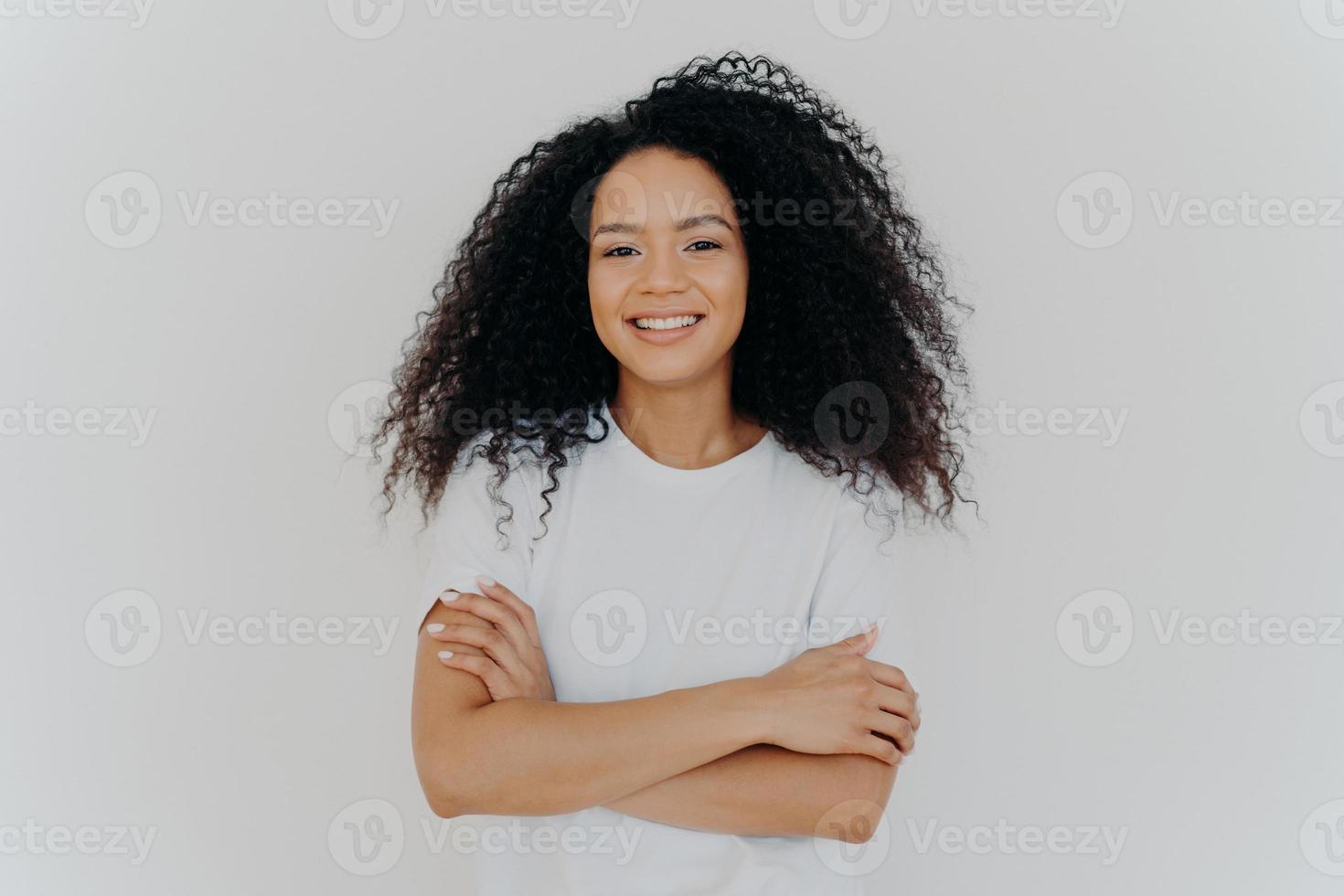 retrato de una joven mujer de aspecto agradable con cabello rizado, mantiene las manos cruzadas, usa una camiseta blanca, sonríe ampliamente a la cámara, tiene una expresión tímida y tímida, posa en el interior, tiene belleza natural. emociones foto