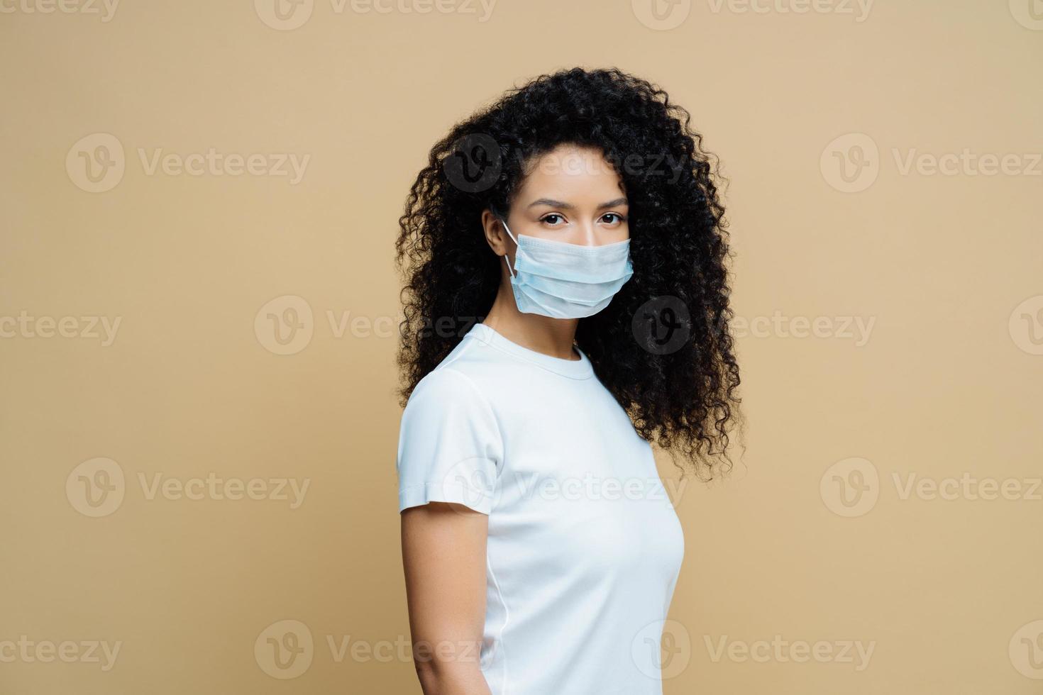 Sideways shot of Afro American woman wears protective face mask, protects against spread of coronavirus disease, stays at home, follows rules of self isolation. Covid-19, safety, quarantine concept photo