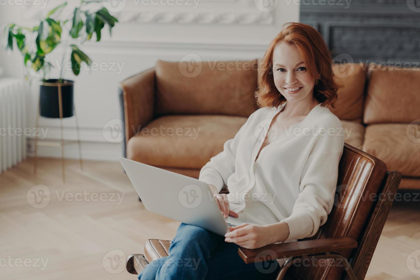 Indoor shot of happy redhead woman freelancer sits at home interior, uses modern laptop computer for distance work, enjoys online communication and free internet connection, waits for answer photo
