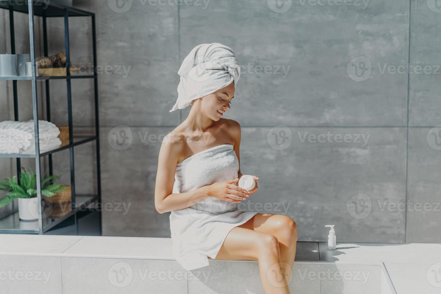 Relaxed young European woman applies moisturizer cream on legs after taking bath sits wrapped in towel in bathroom, enjoys beauty treatments, uses cosmetic product for healthy skin. Hygiene concept photo