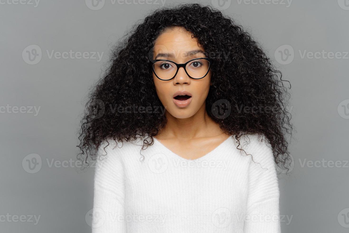 Una foto franca de una mujer hermosa sorprendida con anteojos y cabello rizado oscuro, mantiene la mandíbula caída por el asombro, quedando estupefacta después de descubrir algo sorprendente, aislada sobre un fondo gris