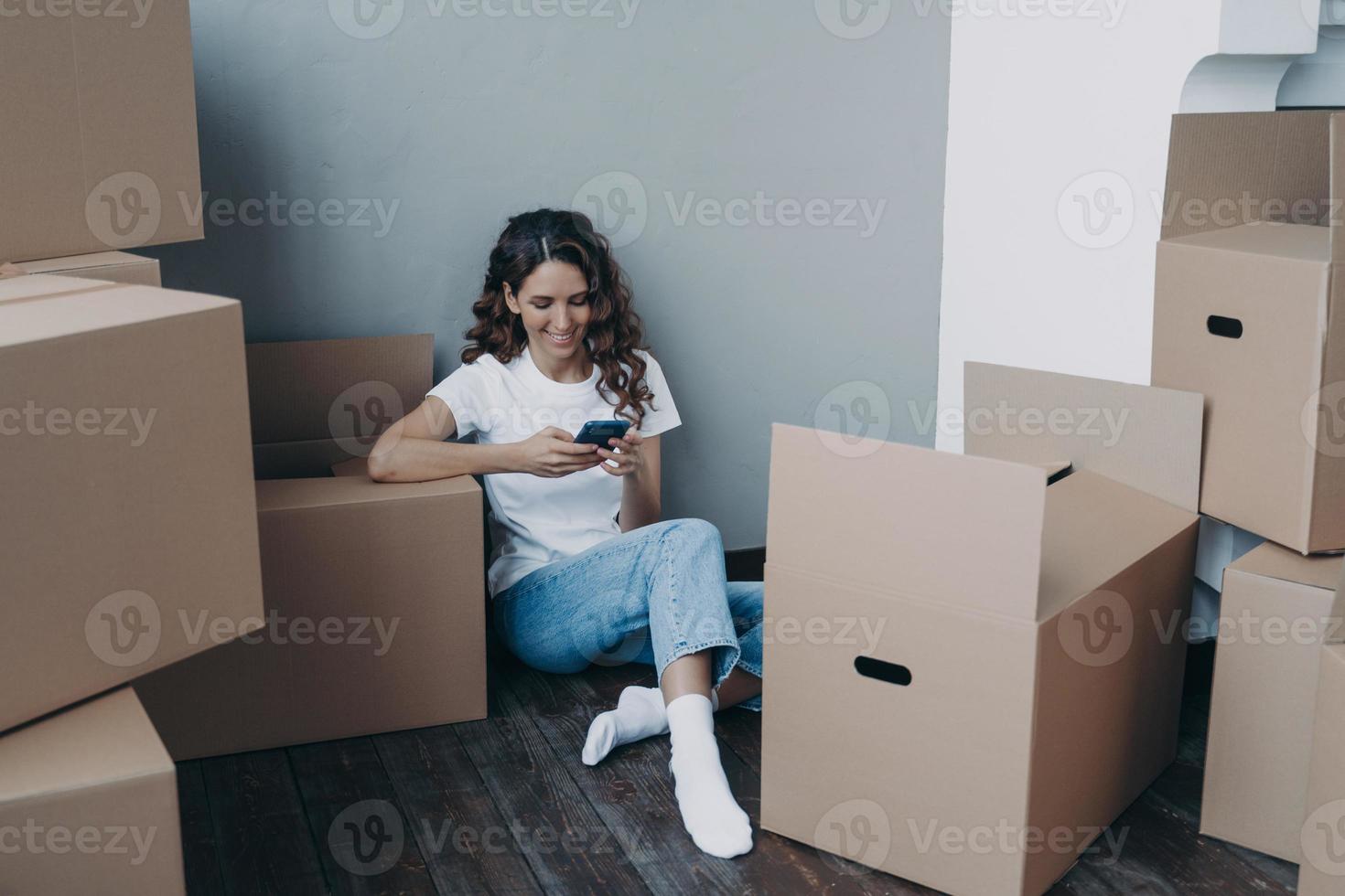 Girl texting on mobile phone and sitting on the floor surrounded with cardboard packages and boxes. photo