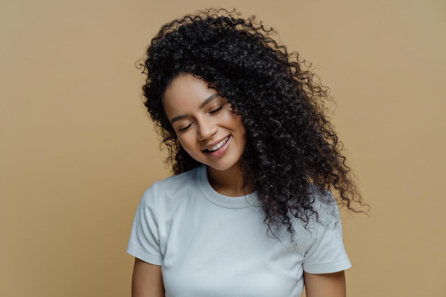 foto de una mujer complacida y satisfecha sonríe con placer, cierra los ojos y muestra dientes blancos como la nieve, tiene un peinado rizado, usa una camiseta informal, posa contra un fondo beige. felicidad, concepto de alegría