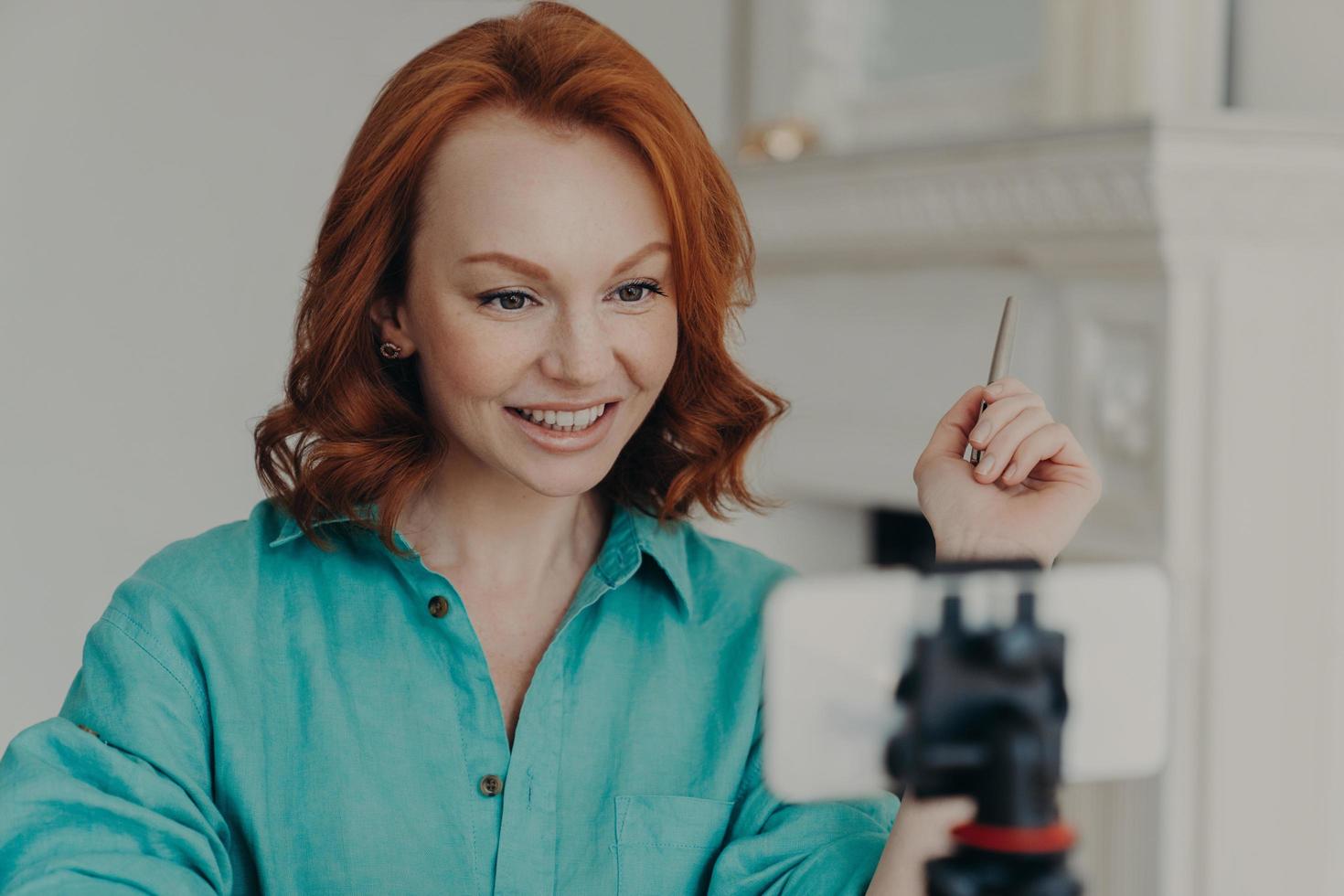 Pleased redhead young European woman uses front cellphone camera on tripod, has personal vlog, shoots video, gives some recommendations, dressed in shirt, talks with followers. Blogging concept. photo