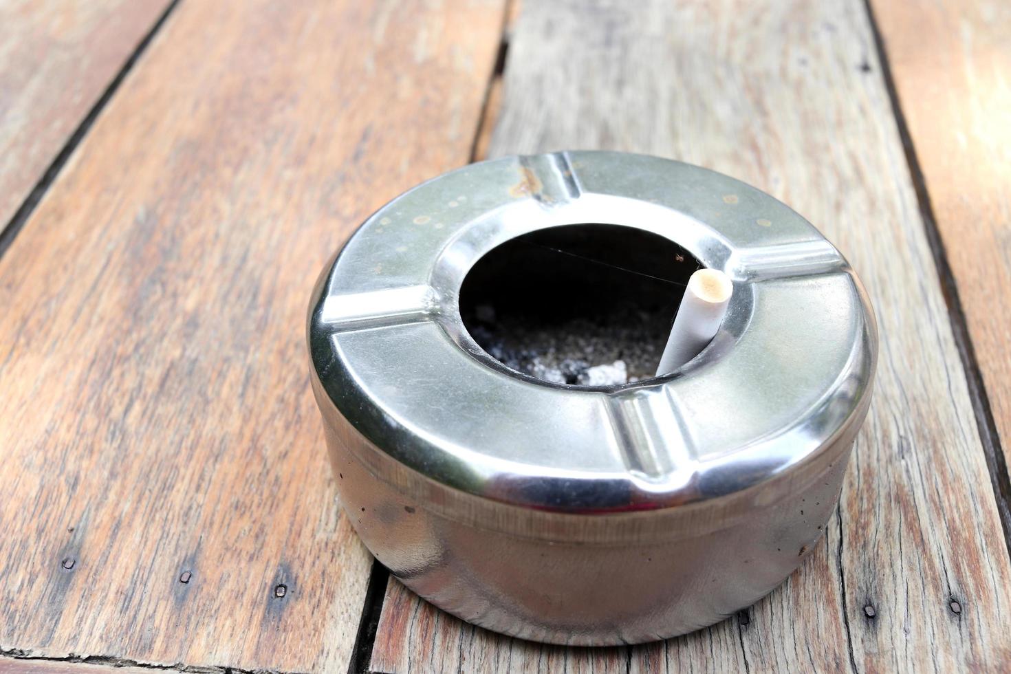 Round shape stainless ashtray is on light brown wooden floor and a cigarette in it, Thailand. photo