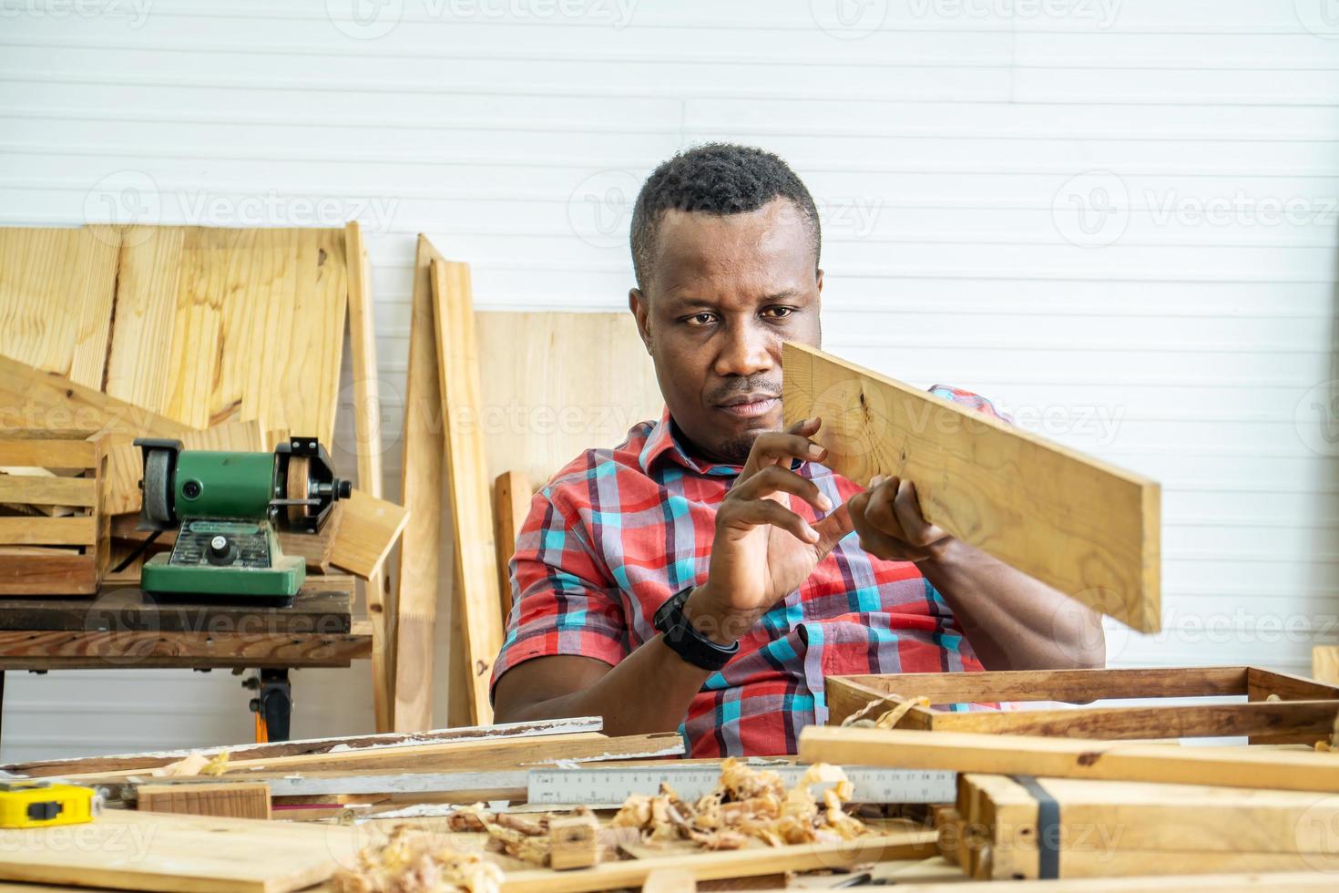 joven carpintero afroamericano mirando y eligiendo madera y usando papel de lija para frotar tablones de madera en la mesa del taller en la fábrica de madera de carpintero foto