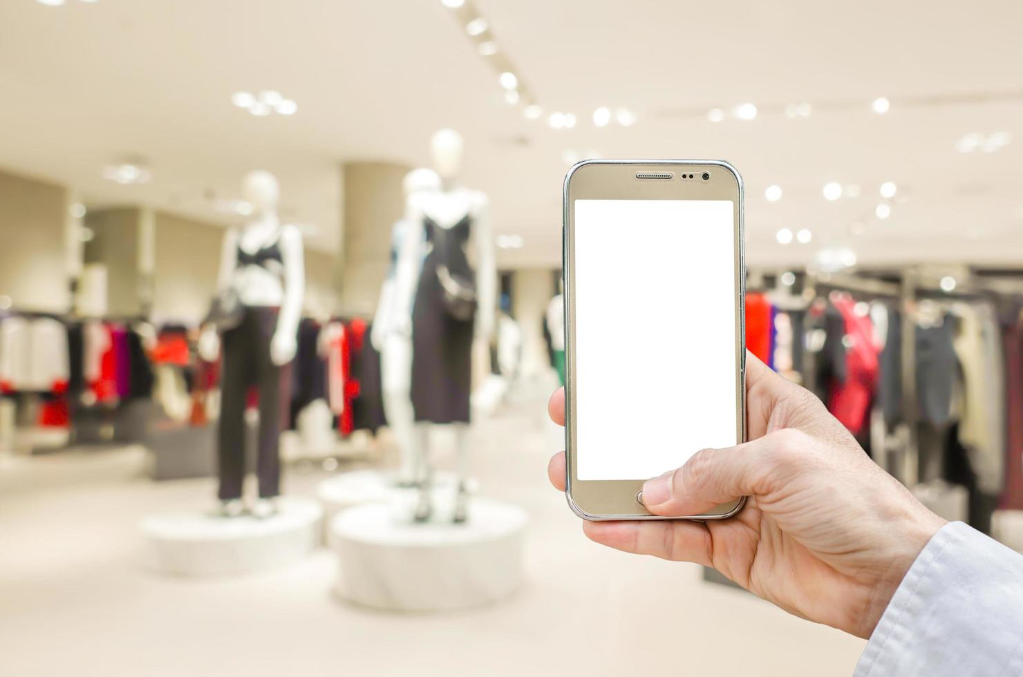 Mockup image of man hand holding gold mobile phone with blank white screen on blurred abstract background of many cotton clothing on the shelves of fashion shop.business concept.shopping online. photo