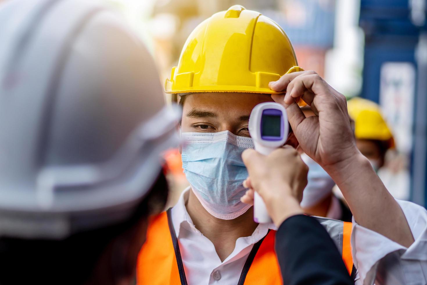 una capataz con una máscara mide la temperatura de un trabajador con traje de seguridad. se pararon y esperaron la medición con un termómetro infrarrojo sin contacto para prevenir el virus covid foto