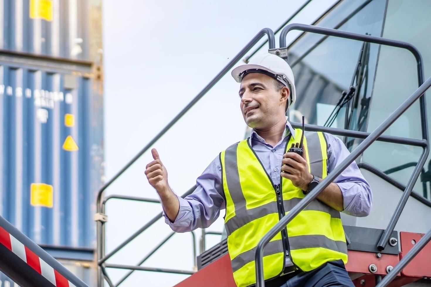 el capataz con casco muestra la señal del pulgar hacia arriba y usa el control de walkie talkie para cargar la caja de contenedores del buque de carga en el concepto de transporte de envío del almacén foto