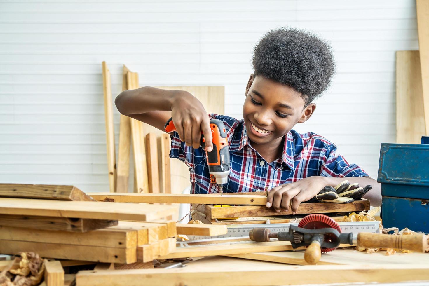 retrato de un niño afroamericano que usa camisa con un taladro en las manos y ayuda a papá a armar un estante de muebles con una herramienta de destornillador eléctrico, concepto de aprendizaje. foto