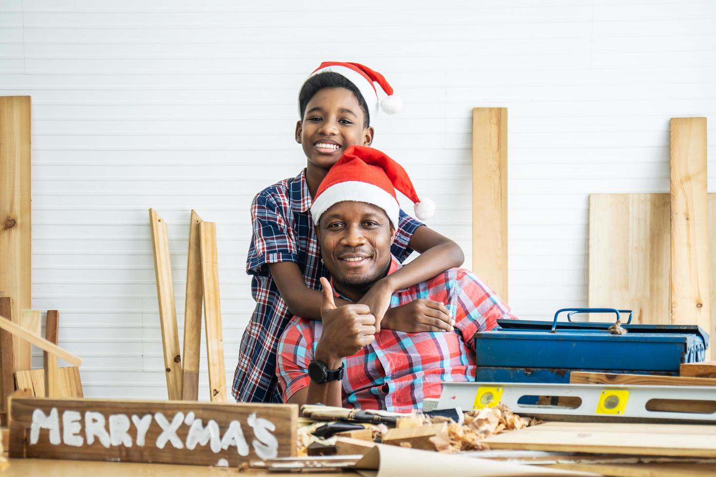 navidad feliz niño y padre. alegre hijo afroamericano carpintero abrazando a su padre mientras se apoya en la mesa de madera con diversas herramientas de trabajo sobre ella foto