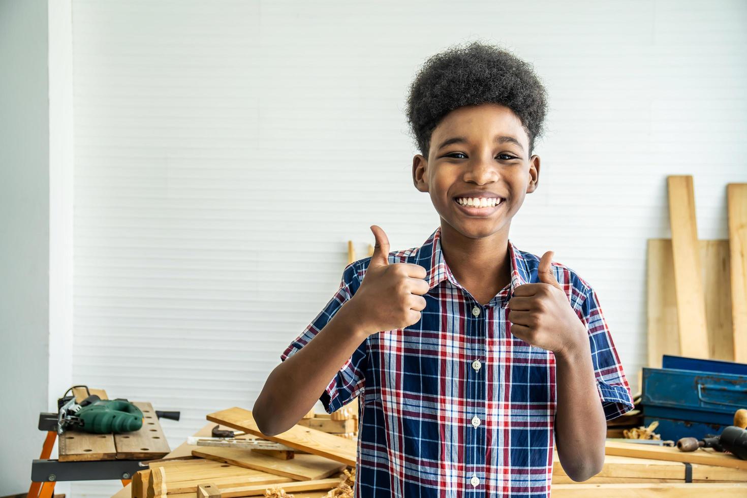 Smiling African-American boy carpenter standing with giving thumbs up as sign of success in a carpentry workshop. photo