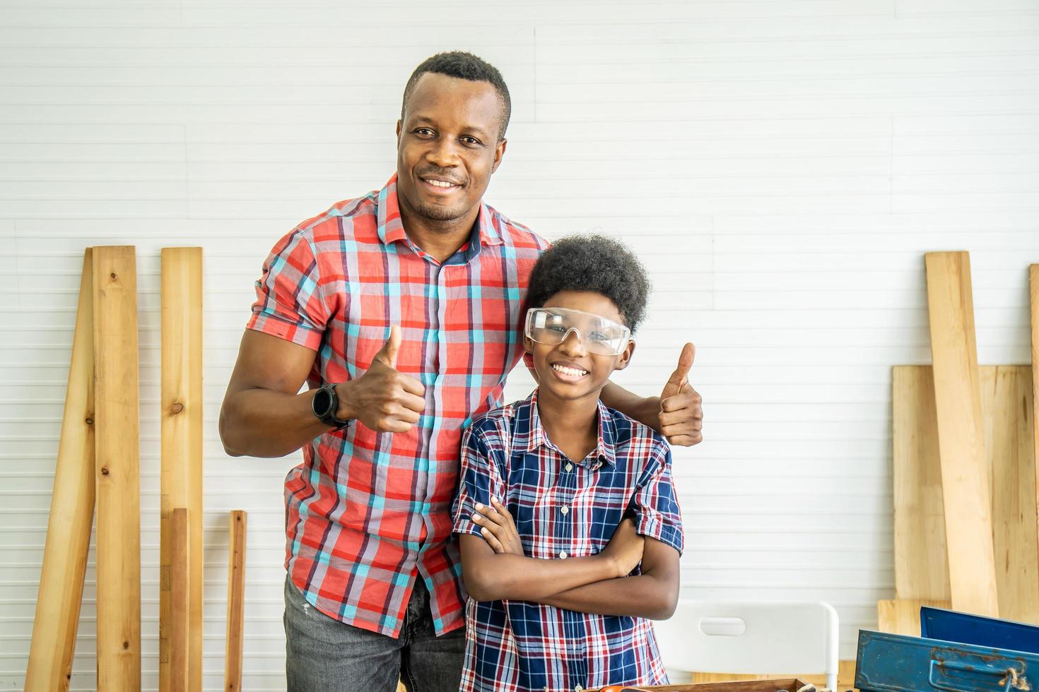family, carpentry, woodwork, Portrait of cheerful african american father and little son making thumbs up at workshop, with diverse working tools laying on it photo