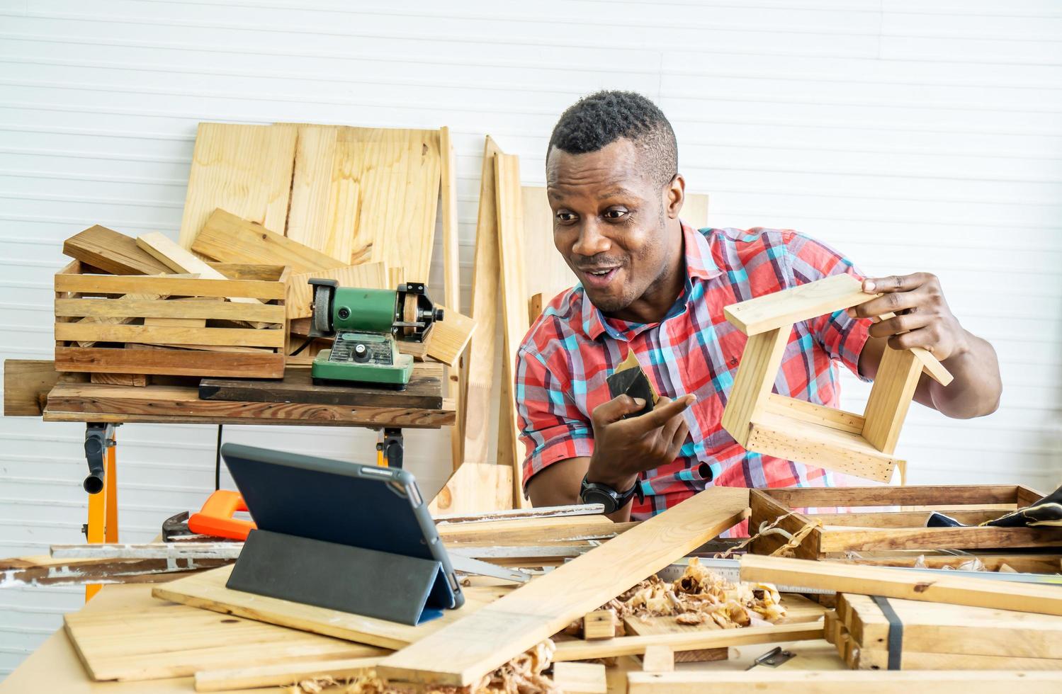 joven carpintero afroamericano sentado en la mesa mostrando artículos de madera a través de una tableta mientras chatea en video en línea con el cliente o enseña en línea en la fábrica foto