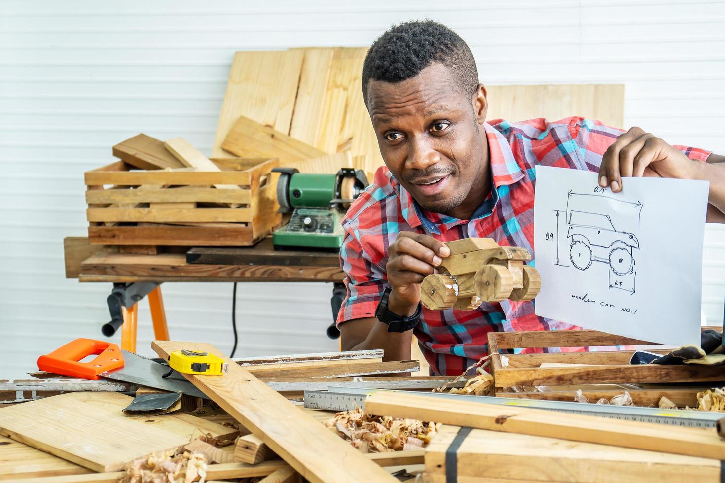 joven carpintero afroamericano sentado en la mesa mostrando modelo de camión de madera y dibujando papel a través de una tableta mientras video en línea chatea con el cliente o enseña en línea en la fábrica foto