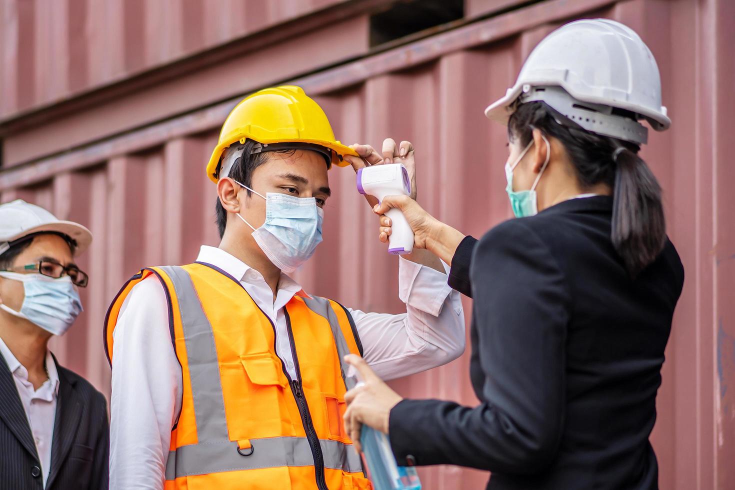 una capataz con una máscara mide la temperatura de un trabajador con traje de seguridad. se pararon y esperaron la medición con un termómetro infrarrojo sin contacto para prevenir el virus covid foto