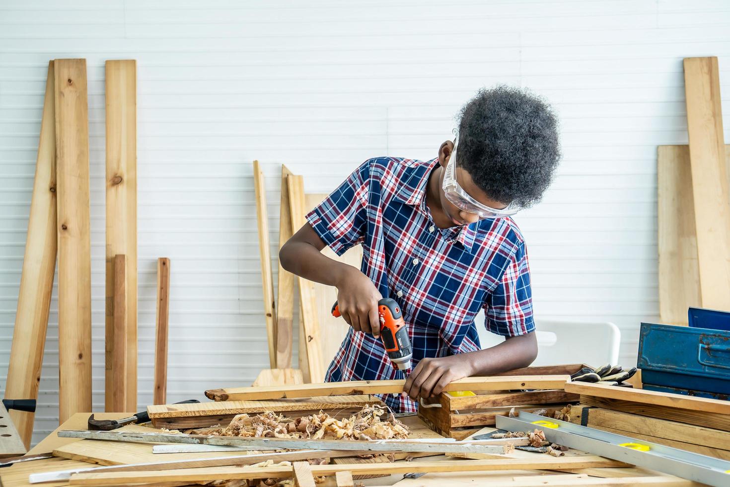 retrato de un niño afroamericano con camisa de pie con un taladro en las manos y ayuda a papá a armar un estante de muebles con una herramienta de destornillador eléctrico, concepto de aprendizaje. foto