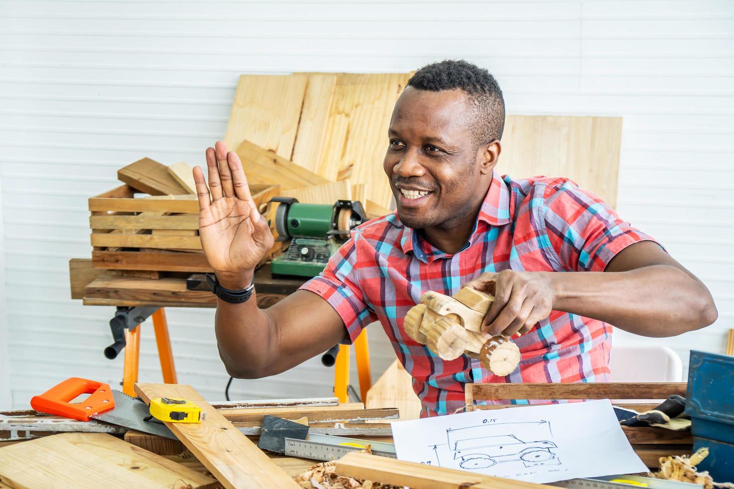 Young african american male carpenter sitting at table showing model car wooden and drawing paper through tablet while video online chatting with customer or teaching online in factory photo