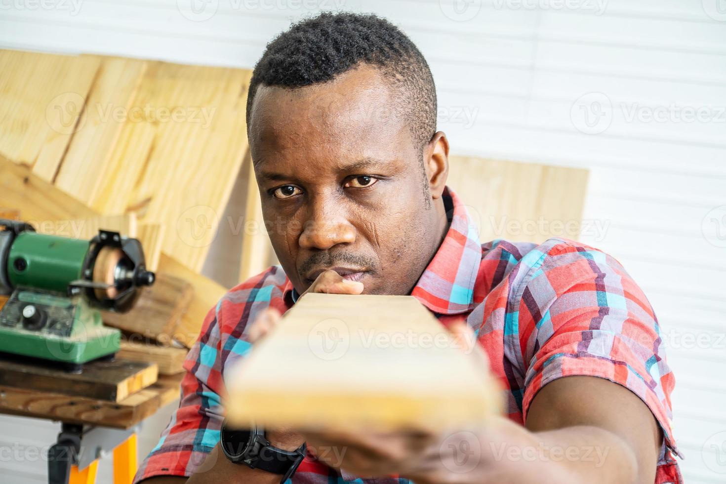 joven carpintero afroamericano mirando y eligiendo madera y usando papel de lija para frotar tablones de madera en la mesa del taller en la fábrica de madera de carpintero foto
