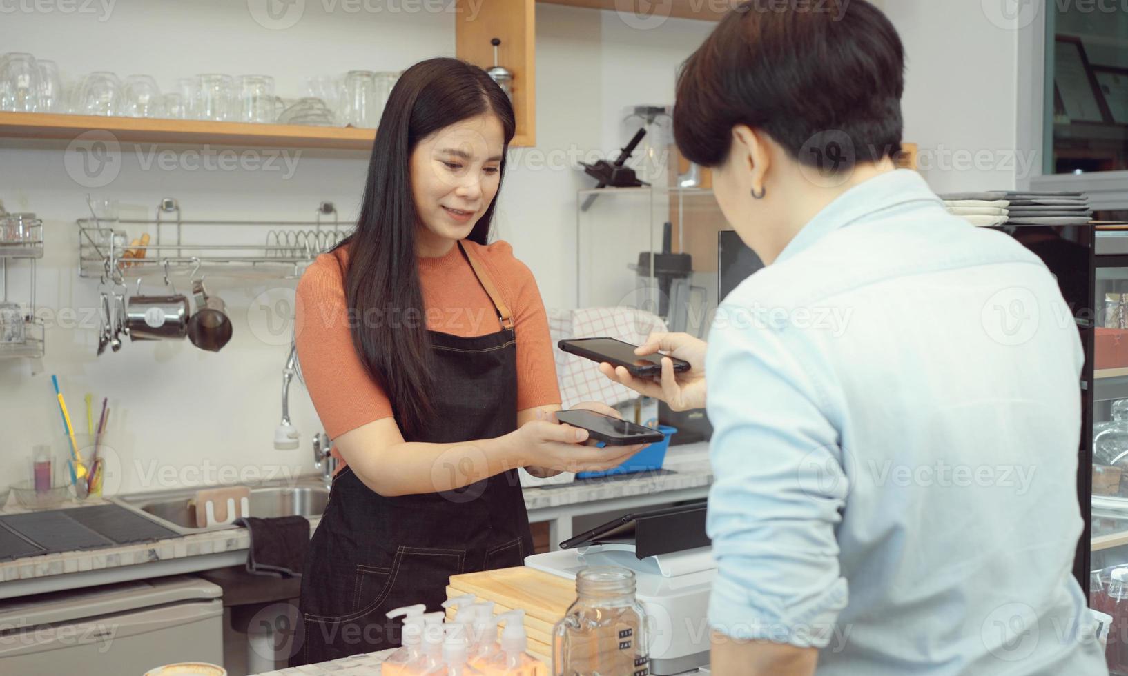 mujer asiática cliente y barista usando un teléfono inteligente para escanear la etiqueta de código qr en otro inteligente con café en una cafetería o restaurante para aceptar generar pago digital sin dinero. foto