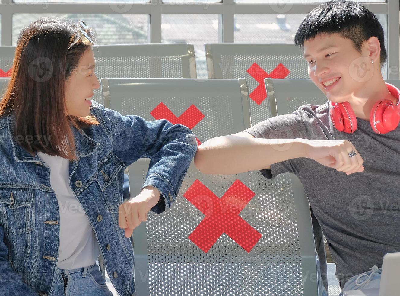 Two asian young traveler with no medical mask arms bump greeting together when meet although following social distancing regulation at public area. COVID-19 infection risk concept. Selective focus. photo