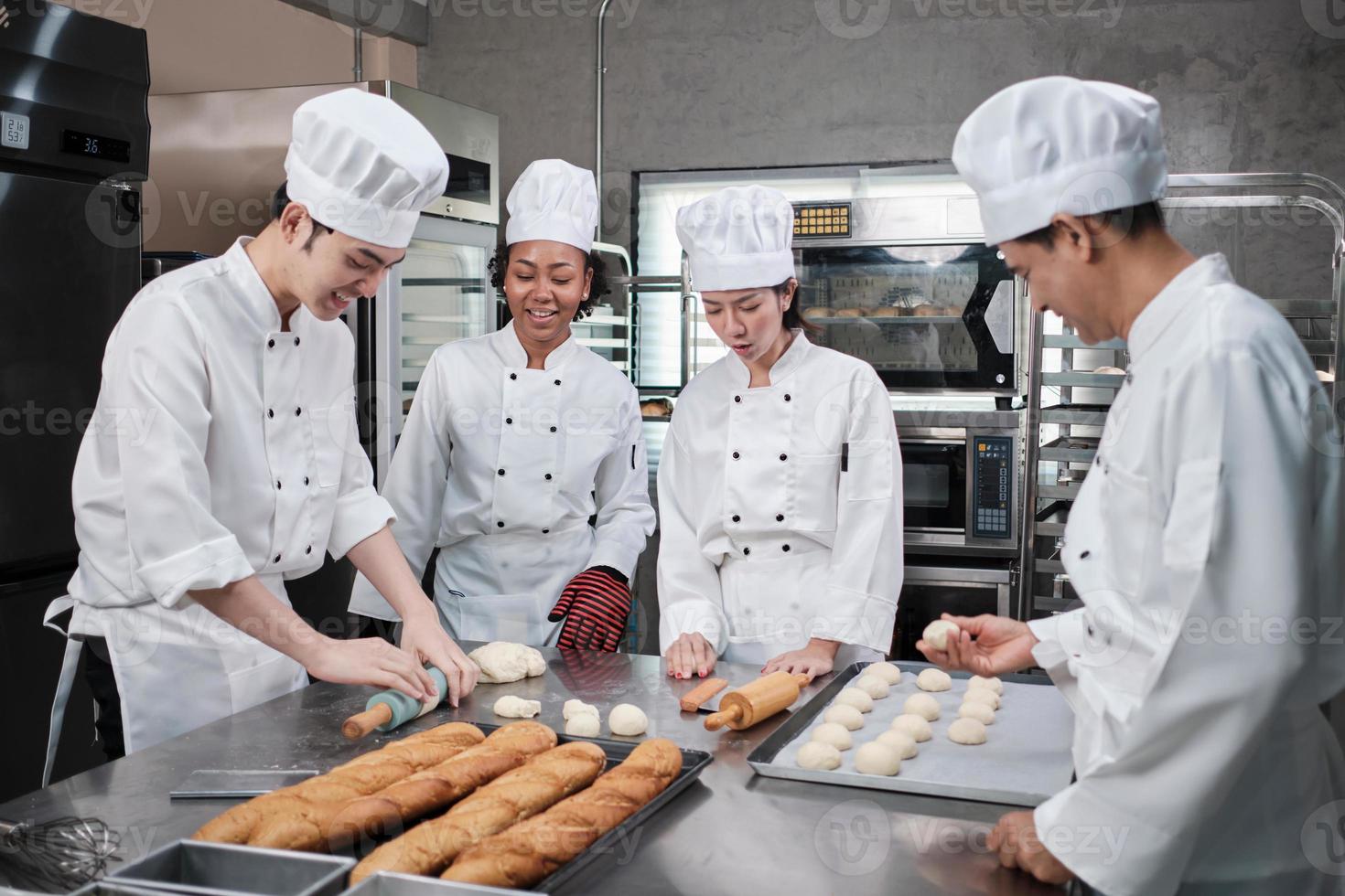 equipo gourmet profesional multirracial, cuatro chefs con uniformes de cocina blancos y delantales amasan masa de pastelería y harina, preparan pan y comida de panadería, hornean en el horno en la cocina del restaurante de acero inoxidable. foto